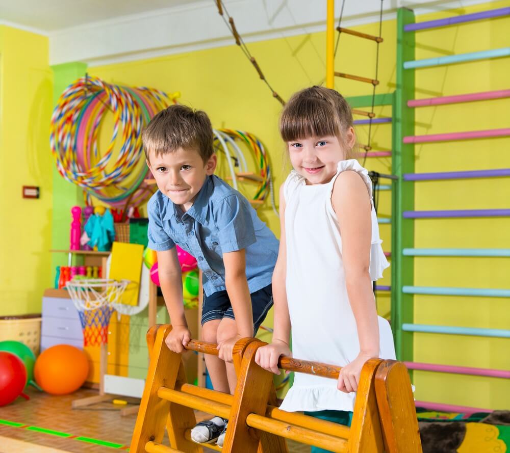 The image shows two young children, a boy and a girl, playing in a colorful indoor gym or playroom. The boy, wearing a blue shirt and dark shorts, and the girl, wearing a white dress, are both smiling and holding onto a wooden climbing frame. The background is filled with various play equipment, including hula hoops, a small basketball hoop, colorful balls, and wall-mounted climbing bars. The atmosphere is bright and cheerful, suggesting a fun and safe environment for kids to play and exercise.